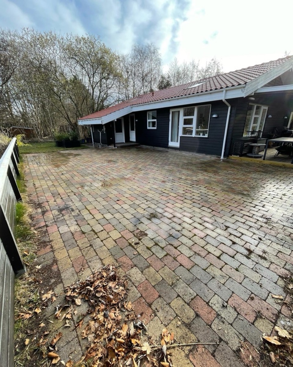 Før: Stor gårdsplads af farvede belægningssten foran et sort træhus